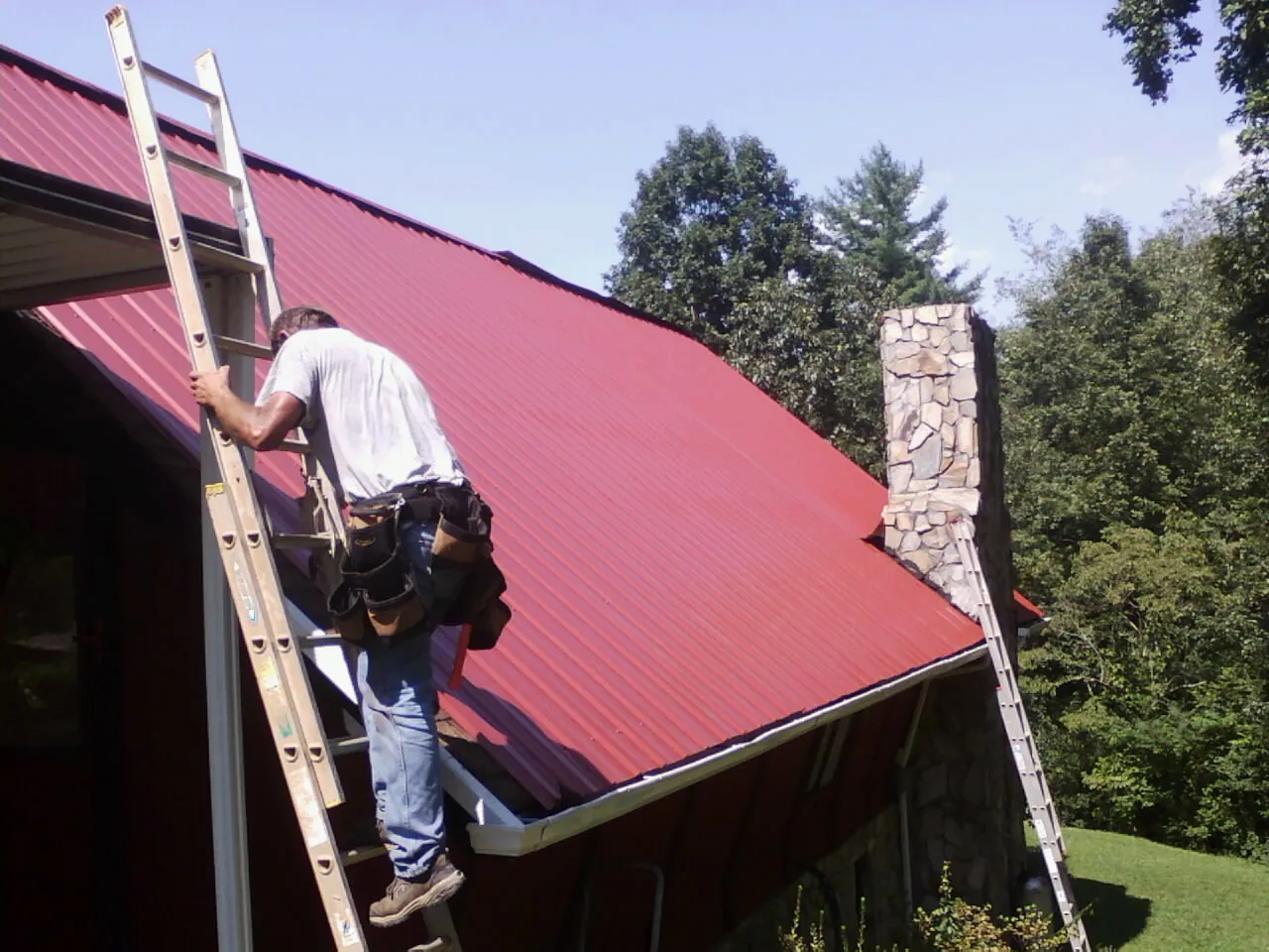 Roofer on a ladder beside a red metal roof and stone chimney — Soffit & Fascia Repair by Roofing Brothers in Stockett